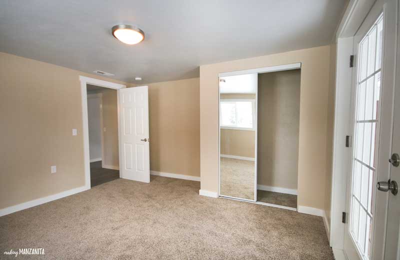 AN image of an small empty bedroom, showing the door and a small walk-in closet with a mirrored door. The room has tan walls and brown carpet.