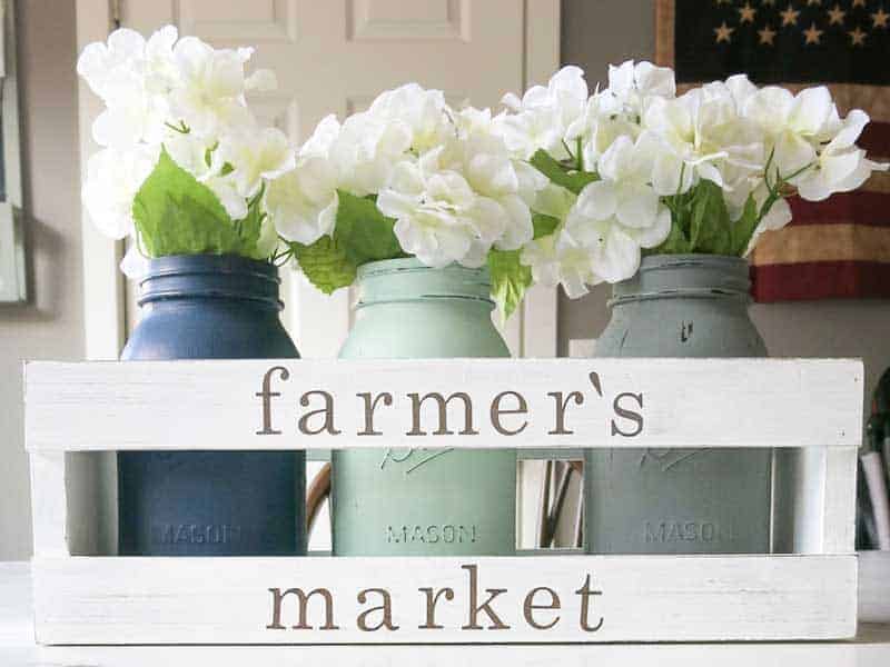 White wooden crate with farmer's market painted on it and three painted mason jars inside with white faux flowers 