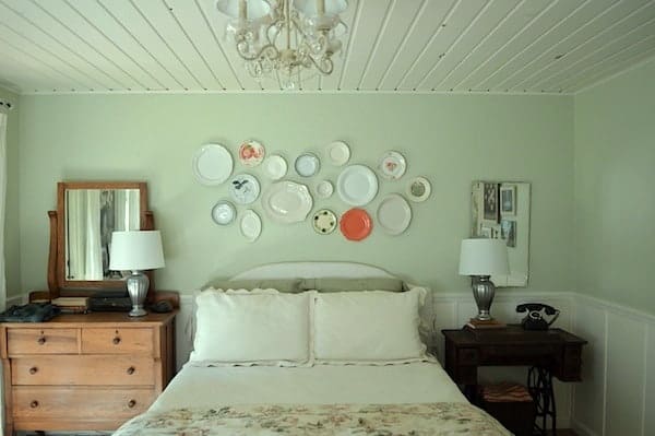 Get creative with the wall decor in your bedroom! This room features a white planked ceiling, white chandelier, vintage plate wall hanging as over the bed decor, white wedding, and dresser used as a nightstand