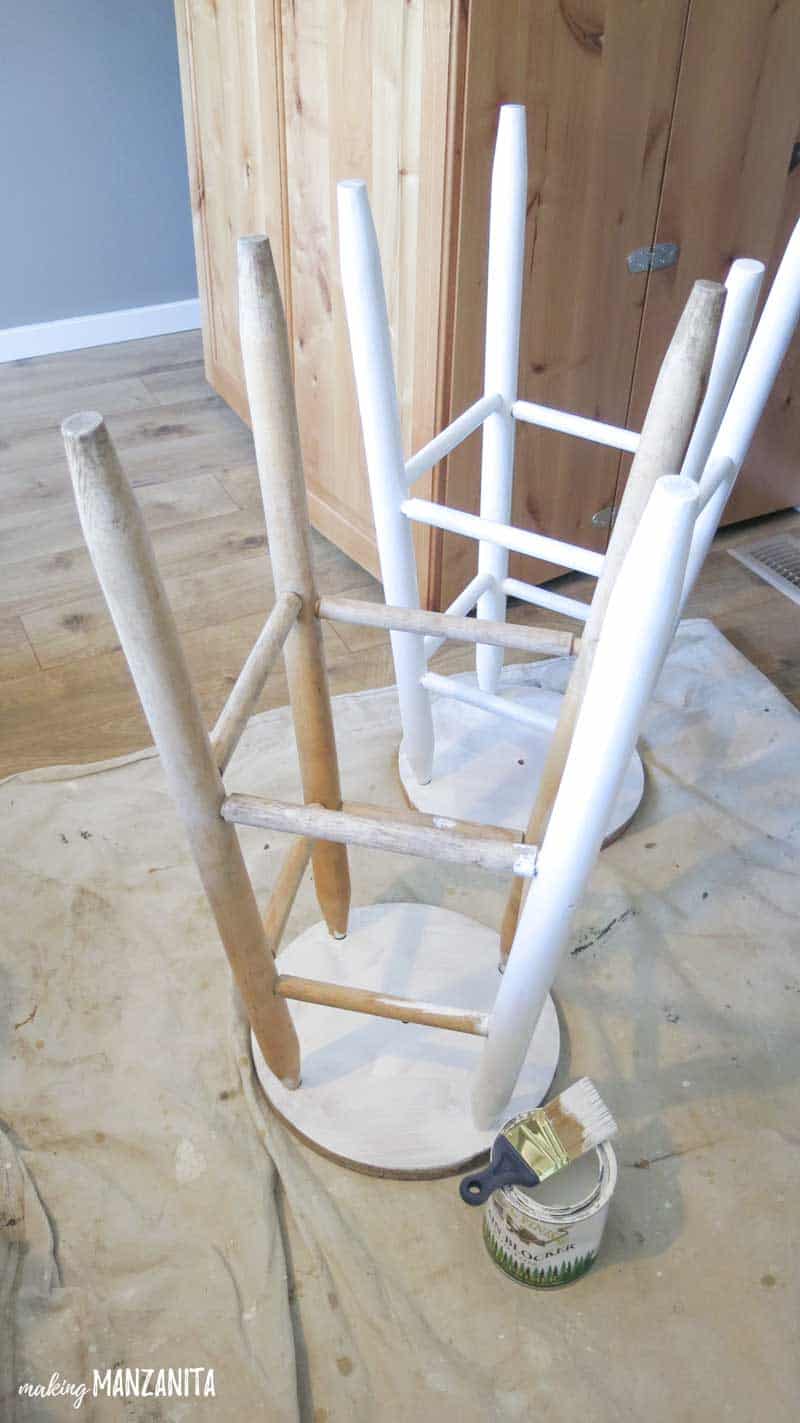 A pair of barstools stand upside down on a drop cloth, in the process of priming with white primer paint.