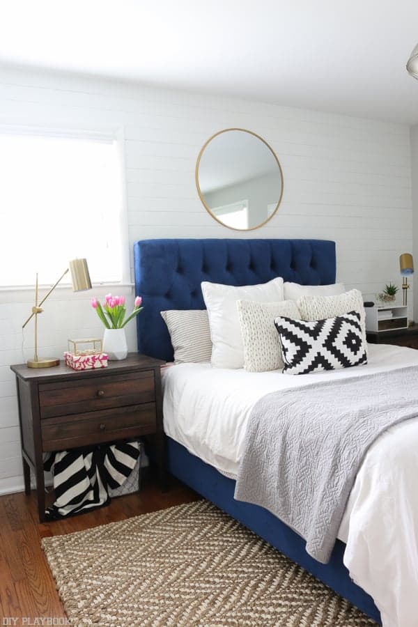 I love this blue upholstered bed with white bedding, jute rug, wood nightstands, white shiplap wall behind bed and gold round mirror hanging as over the bed decor