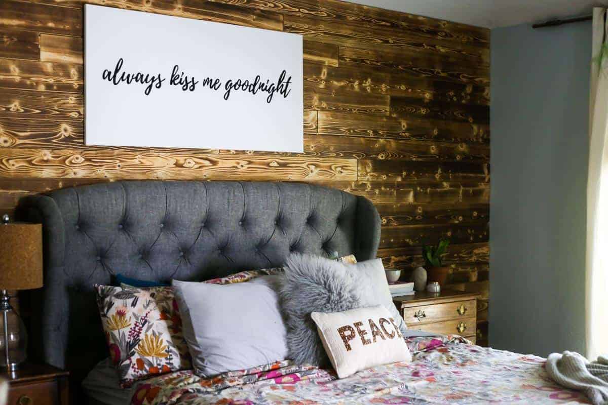 If you're looking for DIY bedroom decor, try a DIY wood planked accent wall. It looks great in this bedroom behind a gray upholstered bed with floral bedding with a sign on wall that says always kiss me goodnight