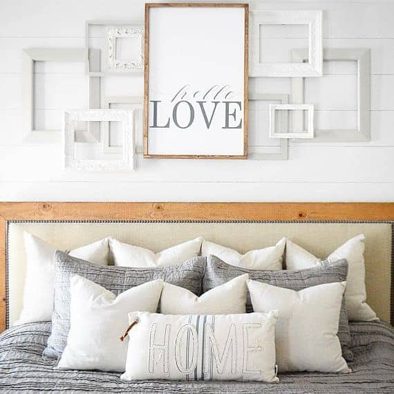 White layered frames and large farmhouse sign are perfect for over the bed decorating ideas. I love this one that that says hello love hanging on a white shiplap wall over a bed with gray and white bedding.