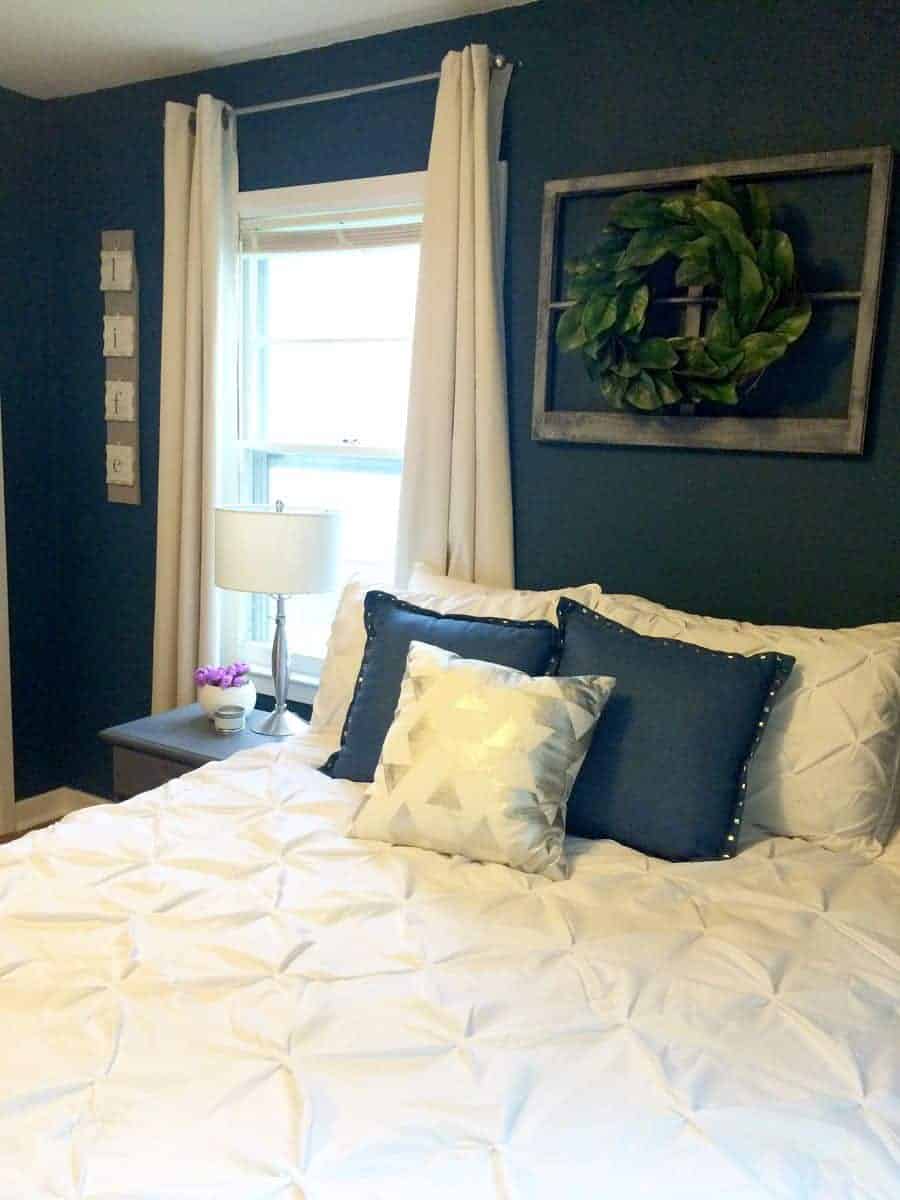 I love this navy painted bedroom with tan curtains, white bedding and a vintage window with a farmhouse magnolia wreath hanging as above bed decor