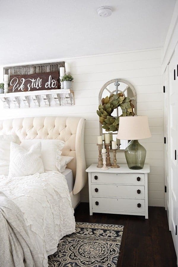 This is such a pretty farmhouse style bedroom with white tufted bed, white bedding, shiplap wall, white painted nightstand, shelf with vintage corbels hanging over bed with a farmhouse sign that says we still do