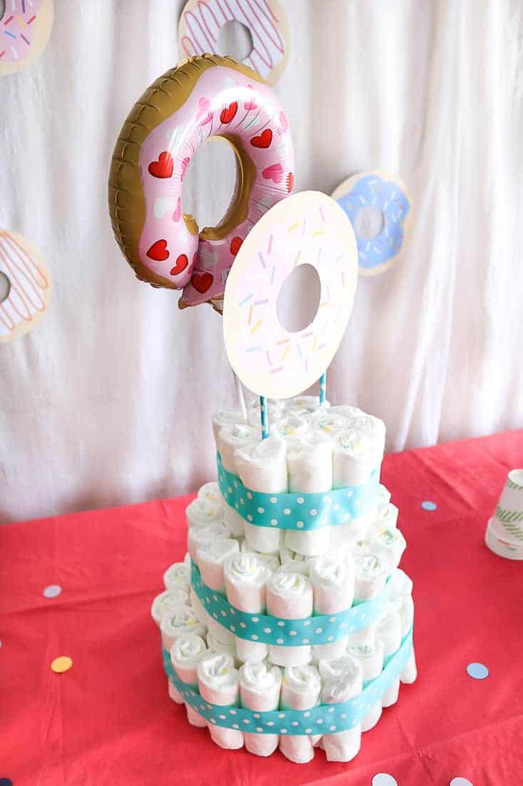 Cake made with nappies with blue and white ribbon accents and donut topper sitting on a table with a bright pink tablecloth