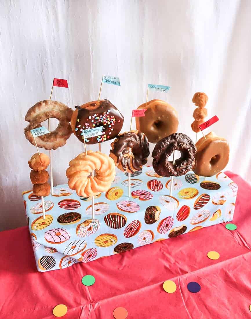 The dessert table at the donut themed baby shower featured a box wrapped with donut print wrapping paper, filled with various kinds of donuts on sticks.
