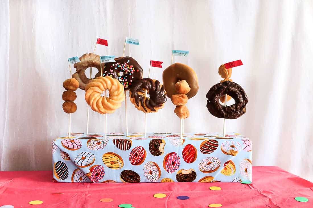 A DIY donut stand, made from a box wrapped in donut print wrapping paper and filled with various kinds of donuts on sticks.