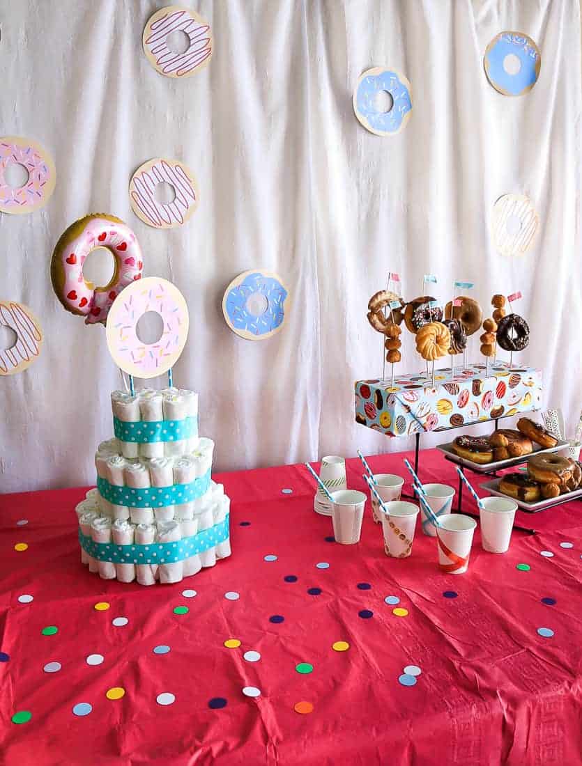 The dessert table at our donut themed baby shower features a diaper cake, donuts and juice, all sitting in front of a white backdrop decorated with donut cut outs.