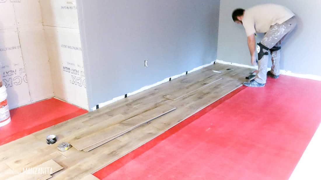 A man installs laminate hardwood flooring over red flooring padding in a large room.