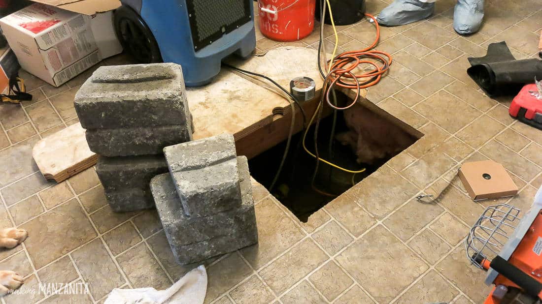 A large hole cut into the floor gives access to a crawl space under the home. There are large bricks stacked next to the hole, surrounded by other construction and home renovation supplies.