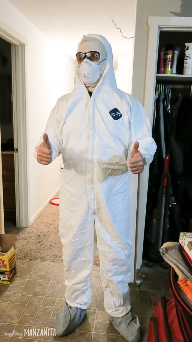 A man dressed in a white protective suit ready to install a vapor barrier under our house in the crawl space. He's wearing eye protection, a respirator mask, and giving two thumbs up to the camera.