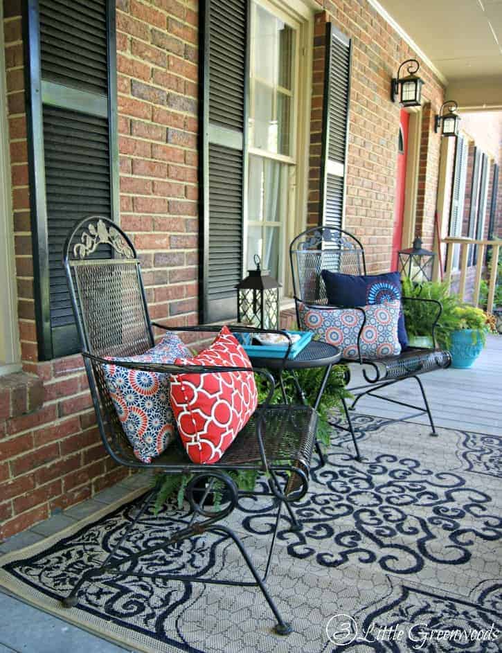 Black metal rocking hairs are decorated with colorful patterned lumbar throw pillows; a matching black and grey rug covers the blue-painted wood porch. 