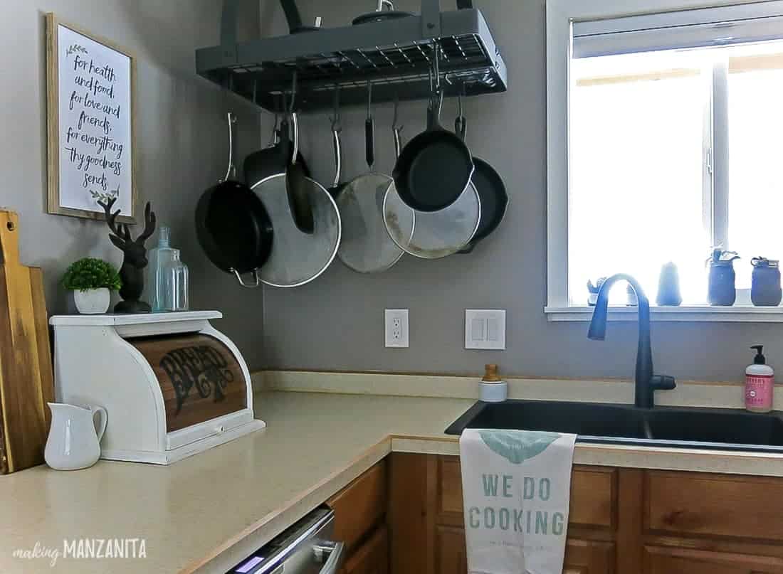 A corner of the kitchen after renovations -- the walls have been painted a light gray, there's a new black metal sink installed, a pot rack, and decor on the countertops.