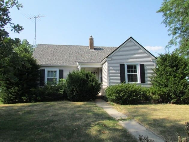 This photo shows a white single-story home with overgrown front yard landscaping and shrubbery covering a majority of the front of the home. 