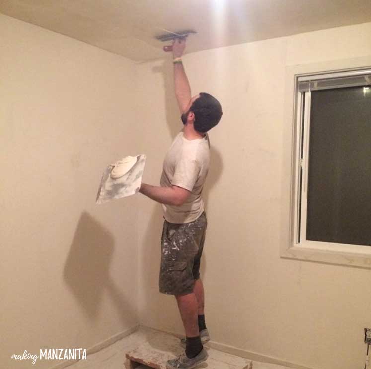 A man applies a layer of skim coating to the ceiling of a white room. He's wearing a white shirt and brown shorts that are splattered with paint.