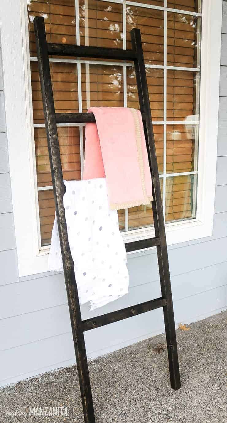 The completed baby blanket latter, fully stained with a dark wood stain, and two  white and pink baby blankets hanging on the rungs of the ladder.