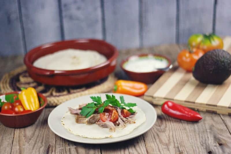 Taco meal shown on colorful plates with peppers, tortillas and avocados in the background. The plates are on top of a pallet wood backdrop perfect for food photography