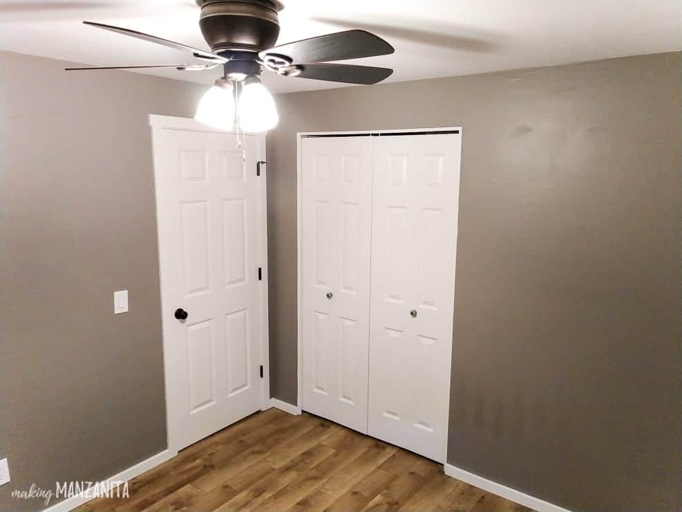 An empty room with gray walls and two white doors -- one entry door, and two folding closet doors. The image also shows the ceiling fan and lights installed on a white ceiling.