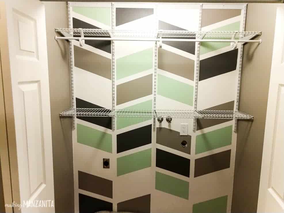 Another hallway closet space, made over into a laundry room with wall-mounted wire shelving and a colorful brown, green, black, and white geometric pattern painted on the wall.