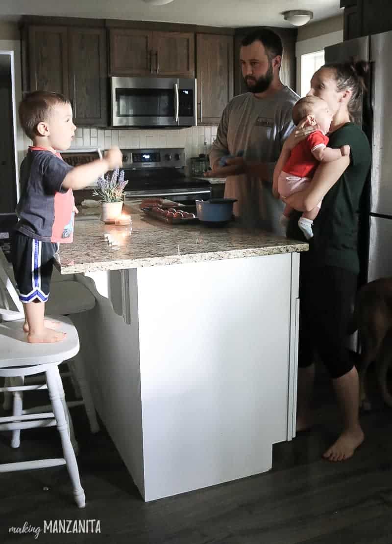 Toddler standing on barstool and kitchen island with parents and baby standing nearby making dinner 