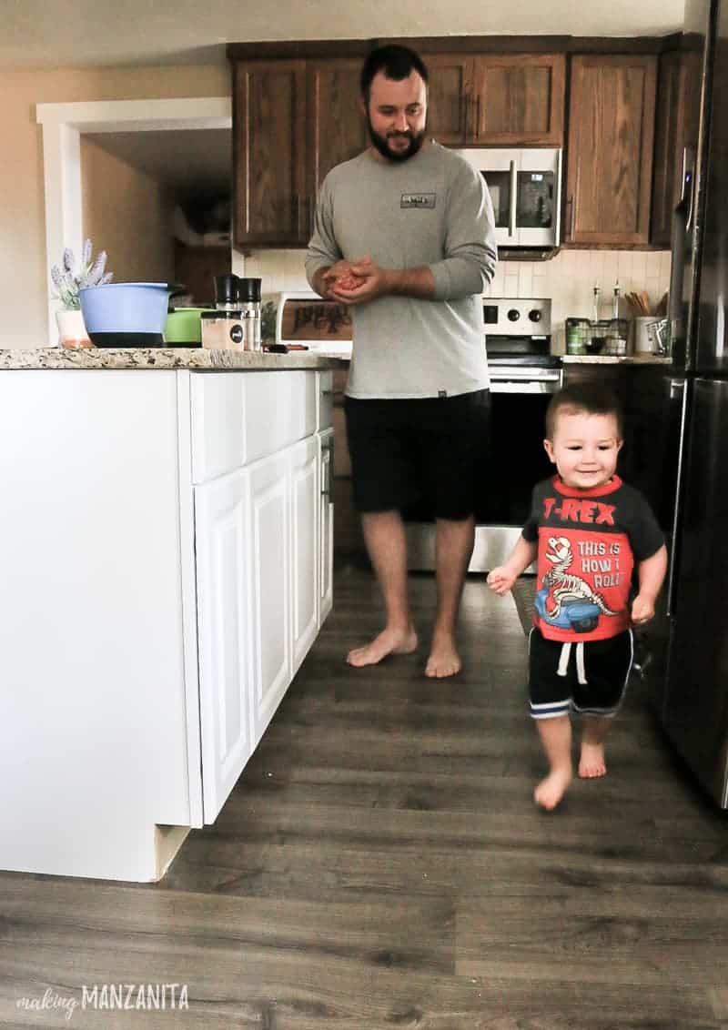Toddler running around kitchen island in kitchen with father in the background cooking 