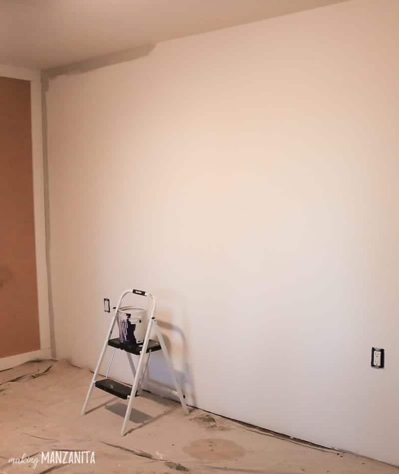 Step stool sitting in front of blank wall  that has been started cutting in with Behr Chic Gray paint color with a drop cloth on the floor with a paint bucket sitting on it