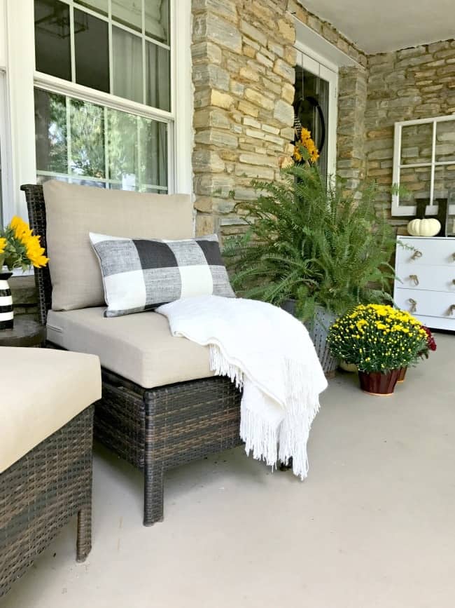This simple fall themed porch is decorated with potted flowers and shrubbery. The porch chairs have tan seat cushions and a plaid throw blanket. The front door is adorned with a simple wreath with yellow sunflowers