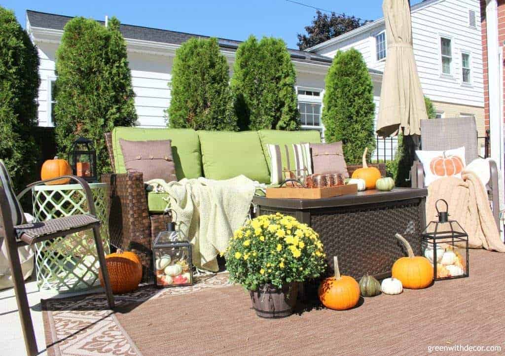An outdoor patio decorated for fall - an outdoor couch has green and brown pillows, pumpkins are scattered across the patio furniture and decorations include blankets, autumn flowers, and lanterns 