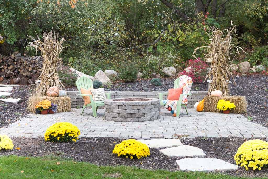 This cozy fall fire pit and sitting area is ready for fall, with haybales decorated with dried corn stalks, yellow and orange flowers, and pumpkins. Green Adirondack chairs sit around the fit pit, decorated with plaid throw blankets and pillows.
