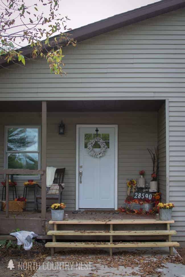 This fall themed front porch is decorated with autumn color flowers and a galvanized metal bucket filled with flowers, faux pumpkins, and vases. The front door is adorned with a simple white wreath.