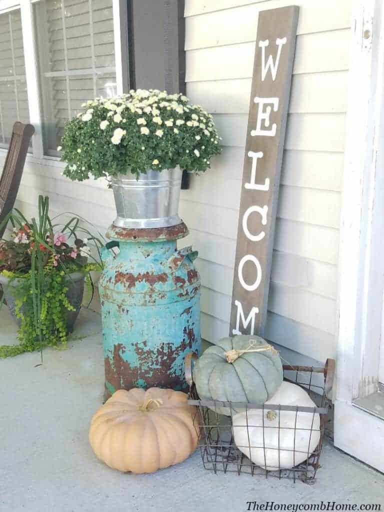 this rustic fall front porch is decorated with a DIY welcome sign, faux pumpkins in a metal basket, and a vintage large metal basin holding white potted flowers.