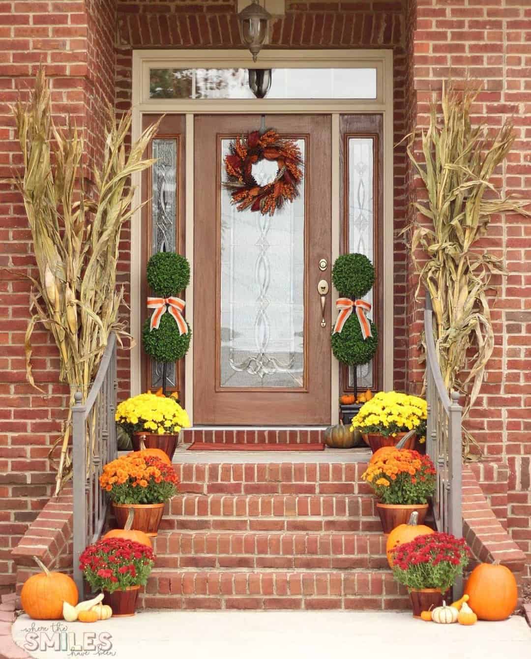 This simple yet traditional fall front porch is decorated with yellow and orange potted flowers, a pair of topiaries with white and orange bows, and dried corn stalks on either side. The front door is adorned with a festive orange and brown wreath.