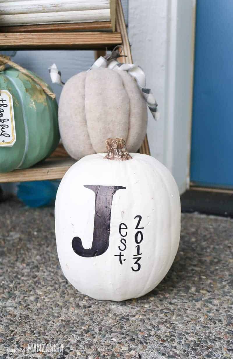 White pumpkin with a big black J written on it sitting on ground in front of wooden ladder with pumpkin decorations. 