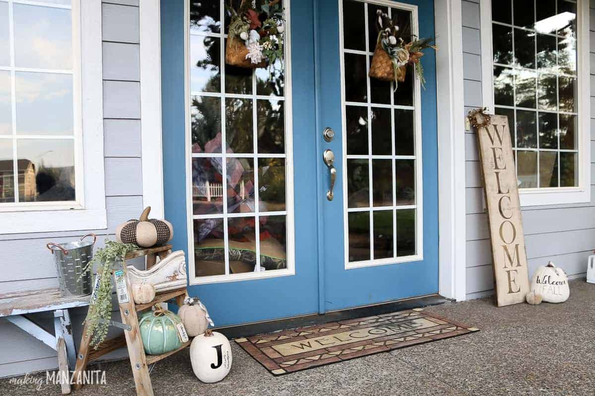 Glass french front doors on front porch with fall decorations including a small wooden ladder with white pumpkin decor, fall door baskets with flowers and wooden welcome sign