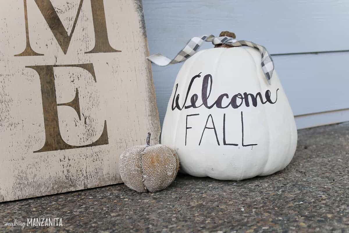 Welcome fall written on white pumpkin decor in black lettering sitting on ground next to large wooden welcome sign