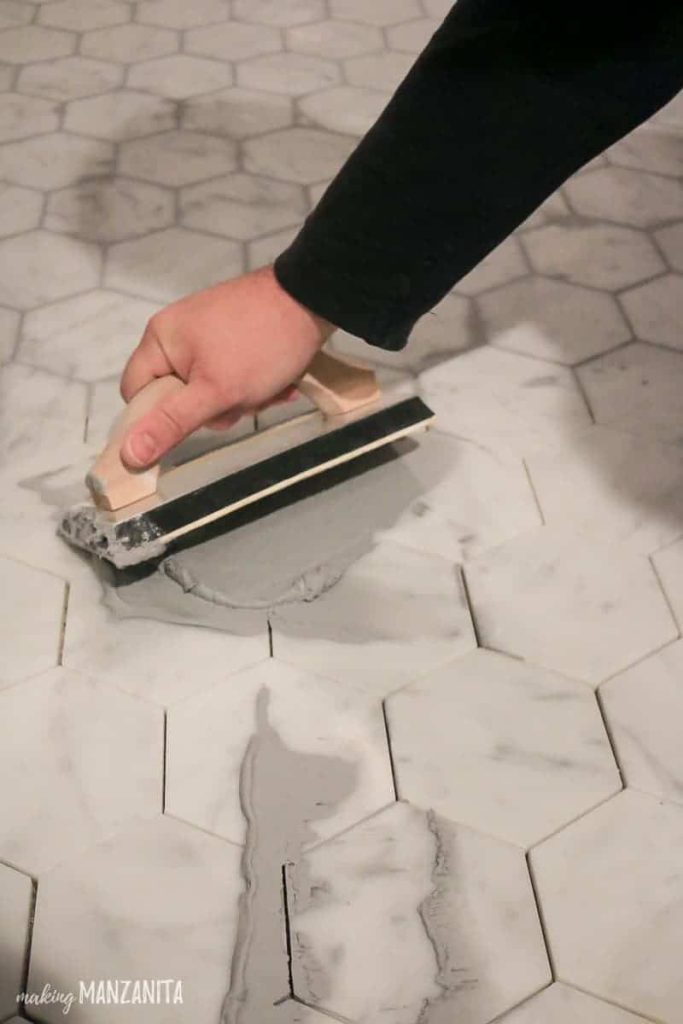 Man applying gray grout over marble tile flooring with rubber float held at a 45 degree angle in a bathroom
