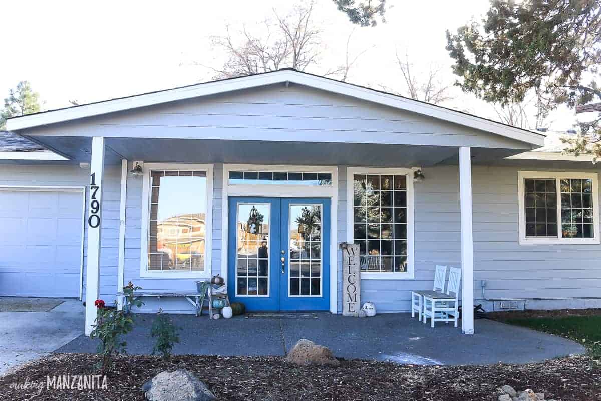 The exterior front entrance to our home - a gable covers a cement patio with front porch decorations, blue french doors. 