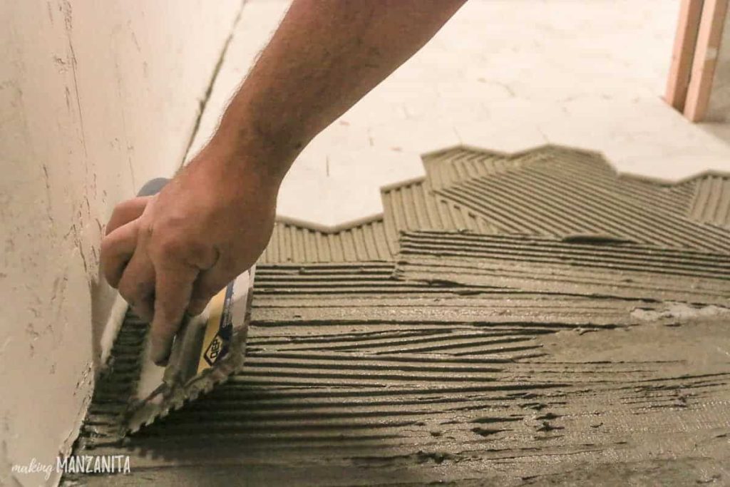 shows a hand using a scraper to put down mortar with white hexagonal tile in the back ground