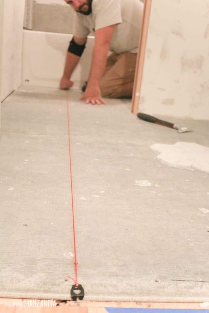 Man sitting on his knees on the other side of the bathroom looking at the concrete flooring with a red chalk line to like up tile placement