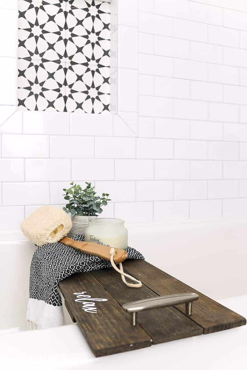 The bath tub in our renovated farmhouse bathroom is accessorized with a wooden bath tub tray sitting in bright white bathtub with subway tile walls and shower niche with background of black and white subway tile