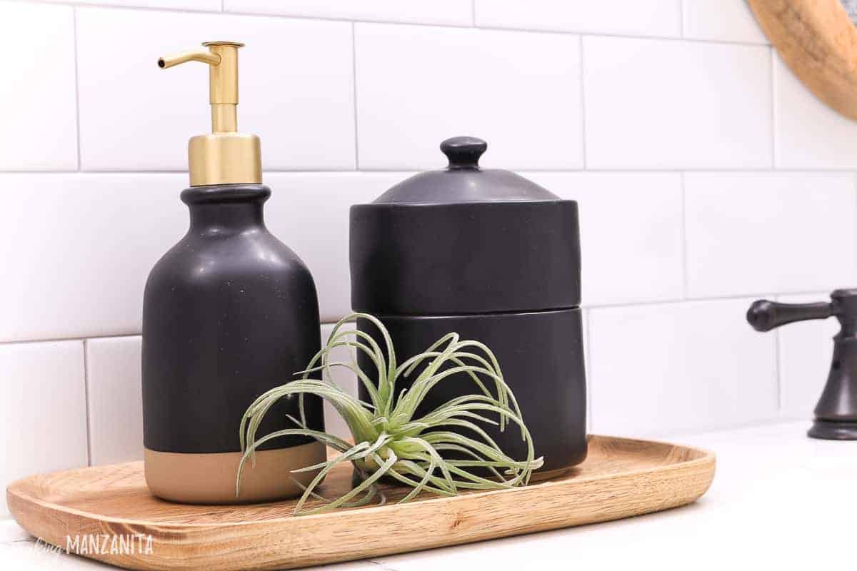 Black farmhouse style bathroom accessories from Magnolia's hearth and hand at Target on countertop with soap dispenser, bath canister to hold cotton balls and Qtips sitting on a wood tray with mini faux airplant