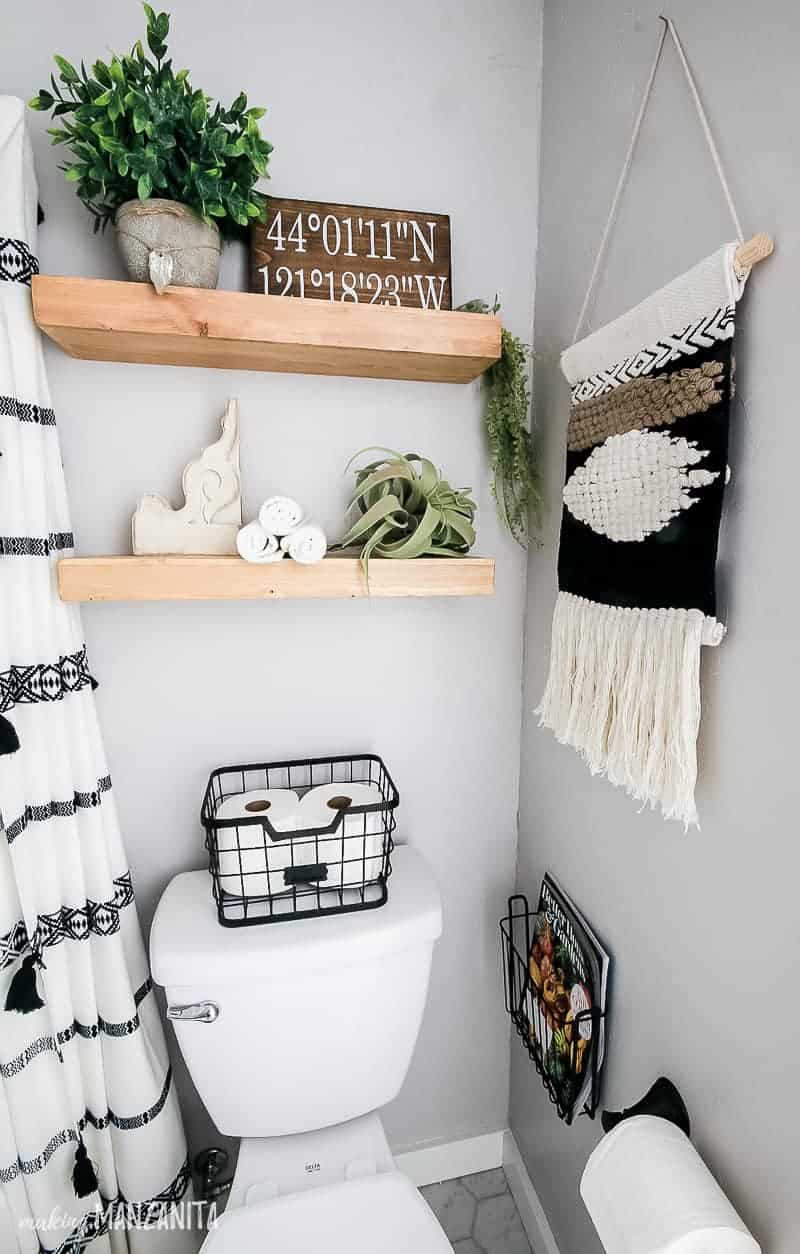 Water closet in our redesigned boho-style bathroom, with light gray walls showing toilet with basket on top to hold toilet paper, magazines, boho woven wall hanging, wood floating shelves with boho styled decor including faux plants, airplant, coordinates sign and wooden corbel