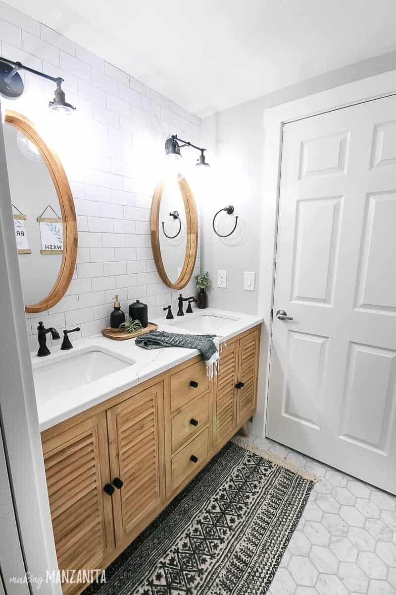 Wood vanity in a boho farmhouse style bathroom with black hardware, dual sinks with white countertop and subway tile backplace with mirrors and industrial style lighting