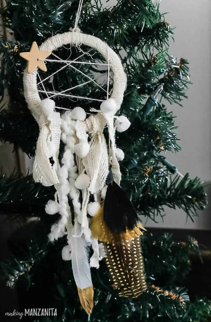 A look at the completed boho style dreamcatcher ornament hanging on a Christmas tree, decorated with hanging muslin fabric, decorative flowers and a boho mini star charm.
