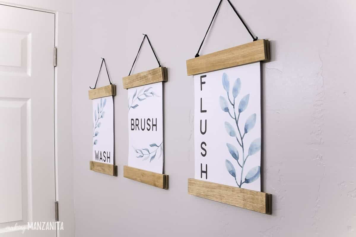 Three printable wall decor pieces hanging in a boho-style farmhouse bathroom that say wash, brush and flush hanging in DIY wood frames 