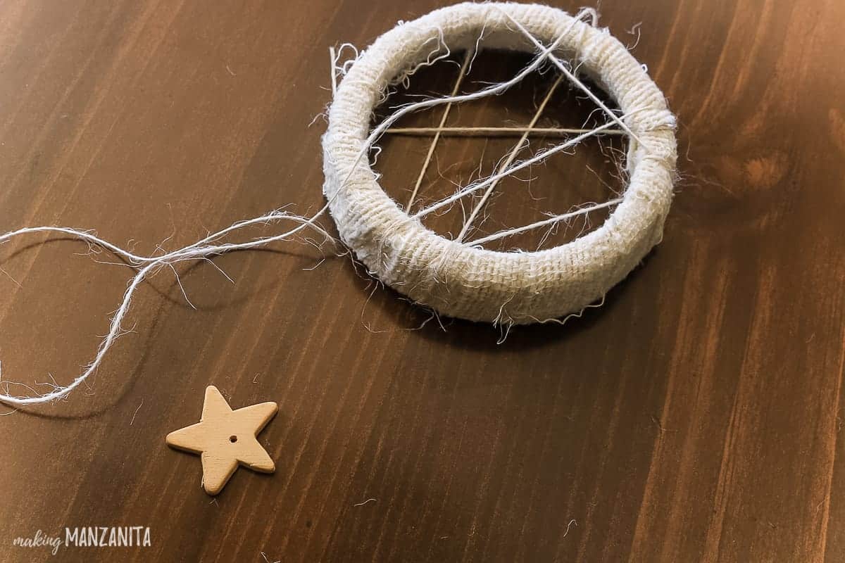 A mason jar lid wrapped in white muslin fabric and craft twine, creating a dreamcatcher ornament, with a small wooden star charm.