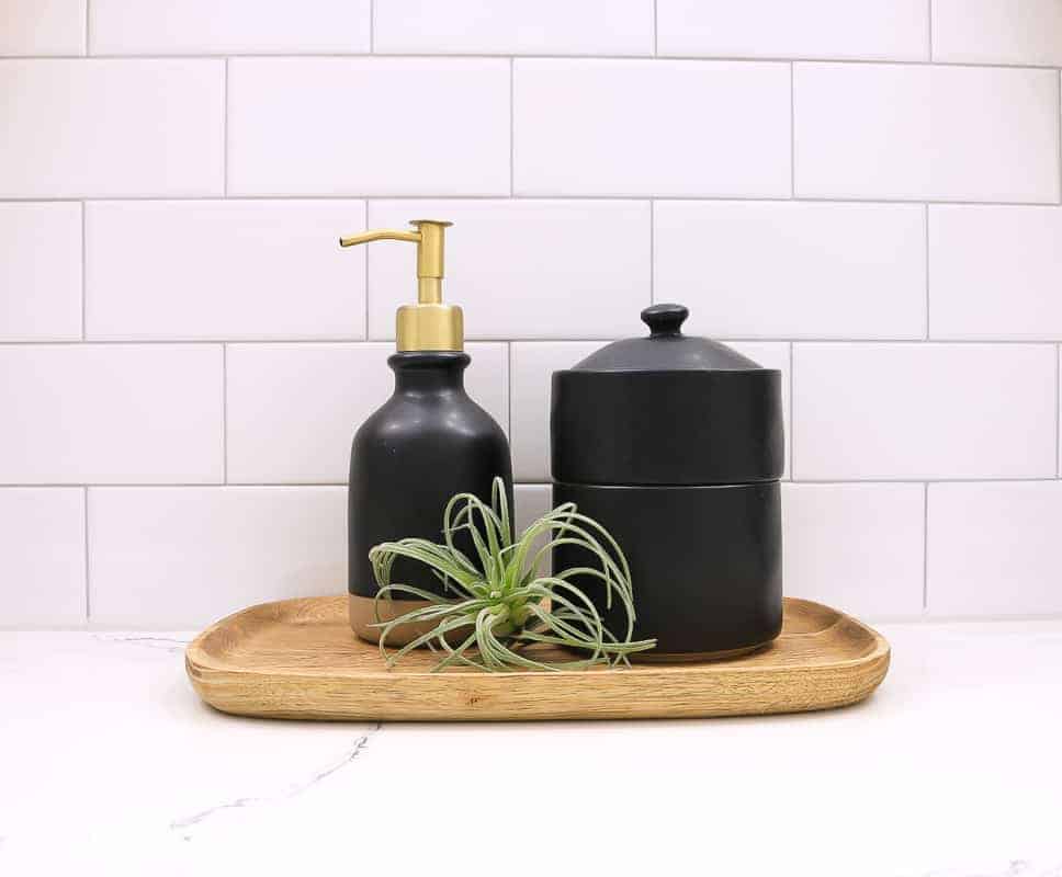 Black soap dispenser and bath canister sitting on wood tray with faux airplant accessorize a boho farmhouse style bathroom renovation