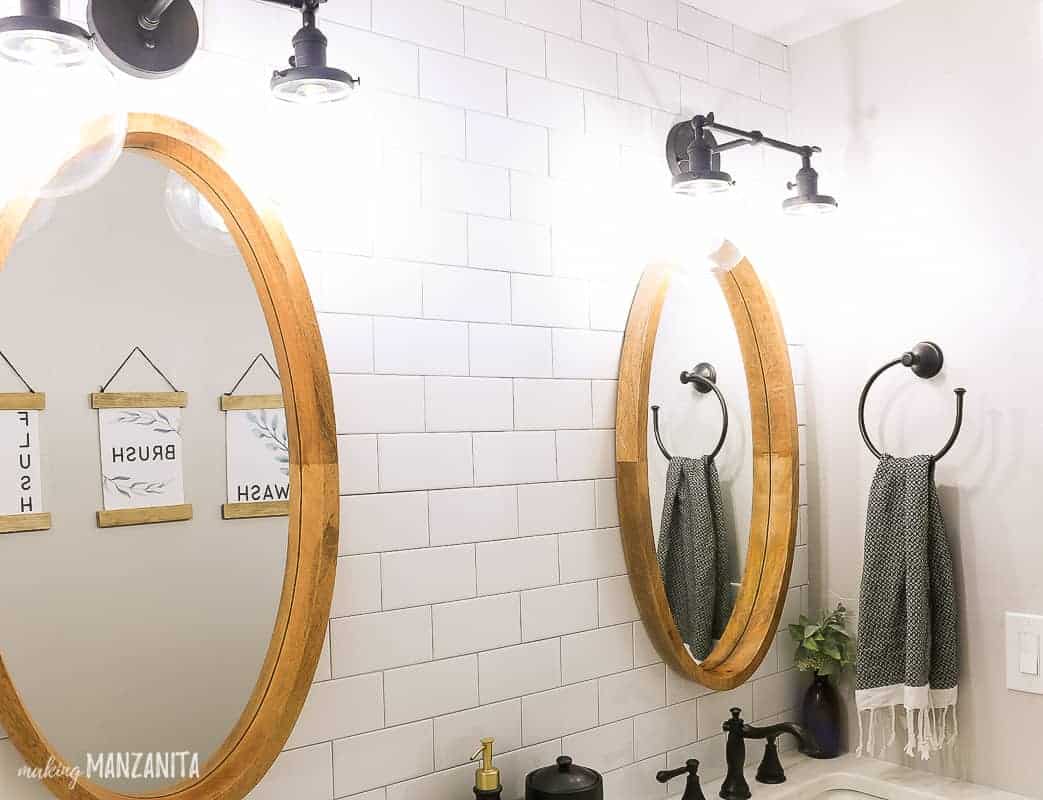 Side by side oval mirror hanging on white subway tile wall with black vanity lights hanging above over a dual sink vanity in a farmhouse bathroom