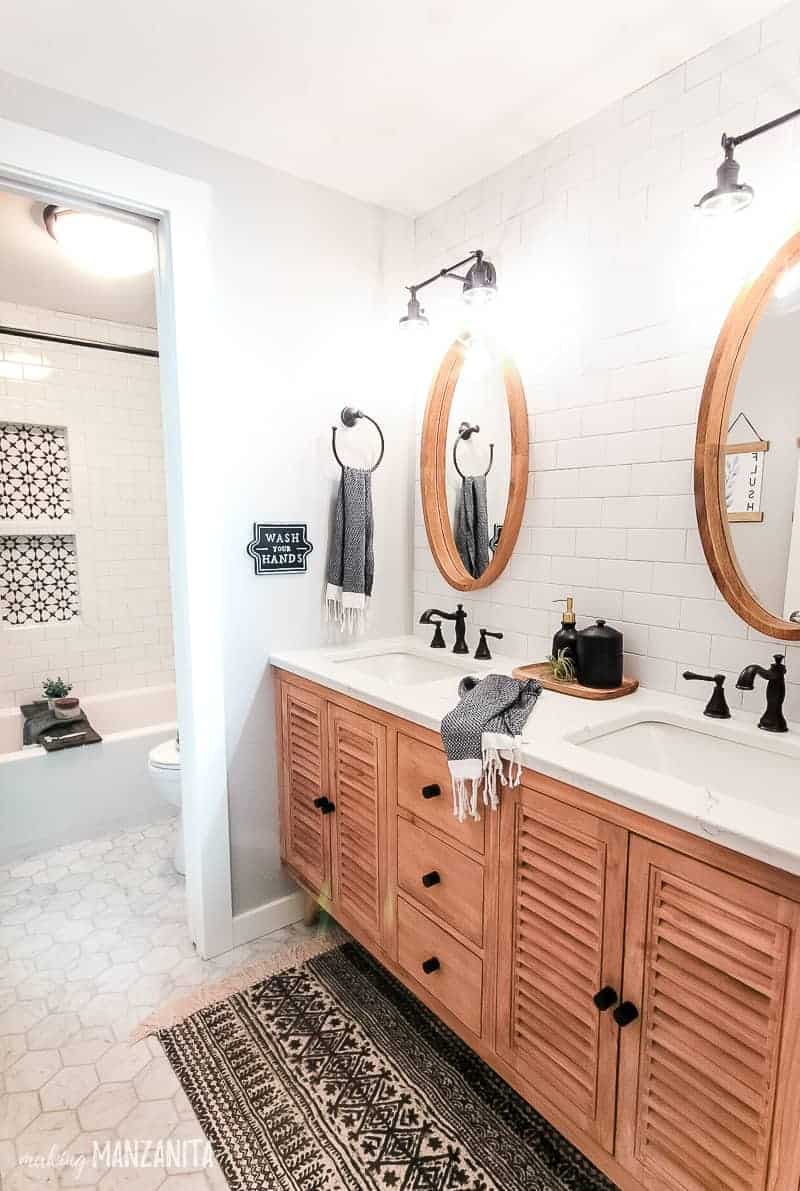 Modern boho farmhouse bathroom with wood vanity and white quartz countertop, gray walls, subway tile wall with round wood mirrors and black vanity lights, the corner of the white subway tile shower is shown in the background